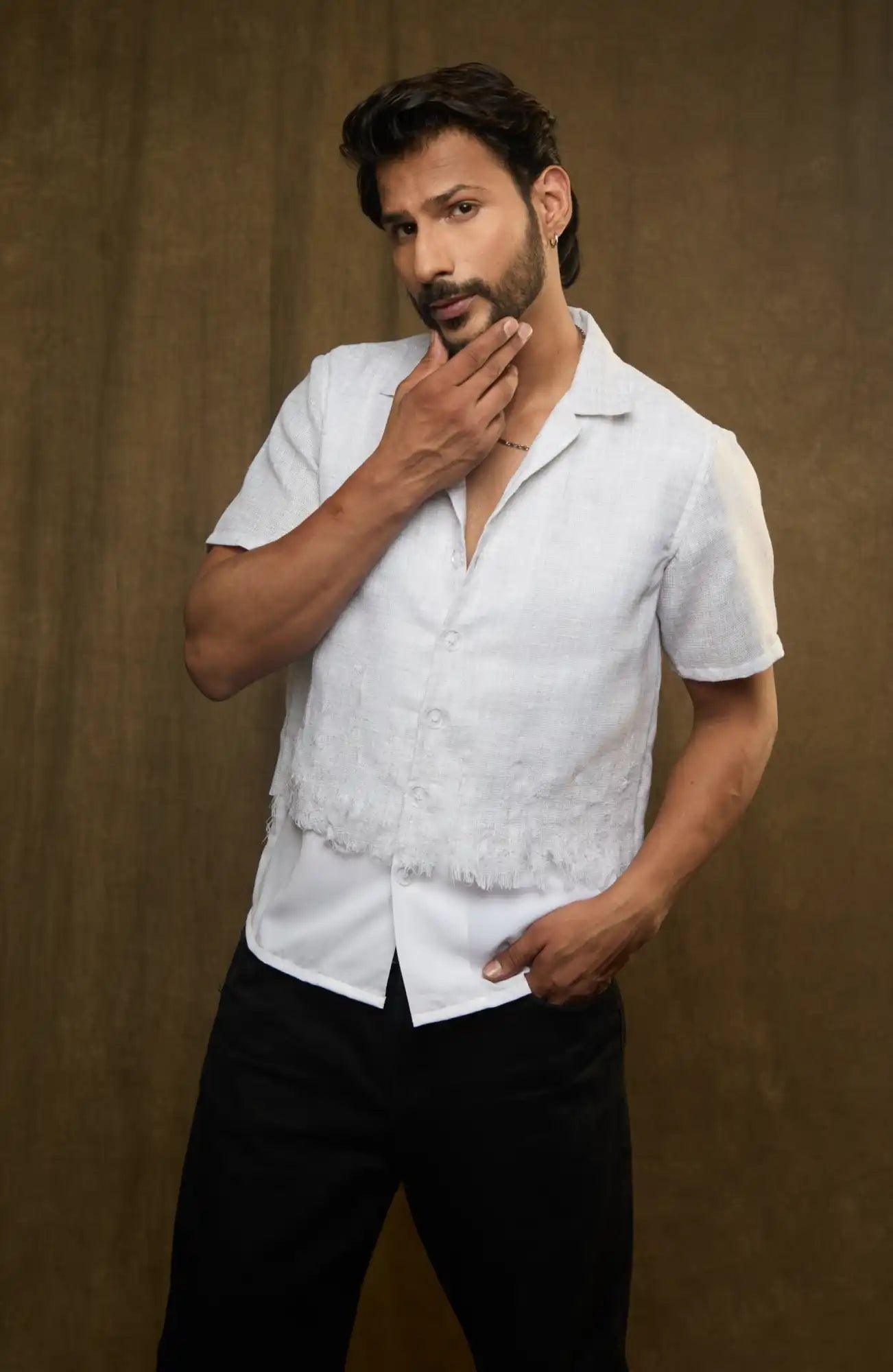 A bearded man wearing a white short-sleeved shirt and black pants in a thoughtful pose.